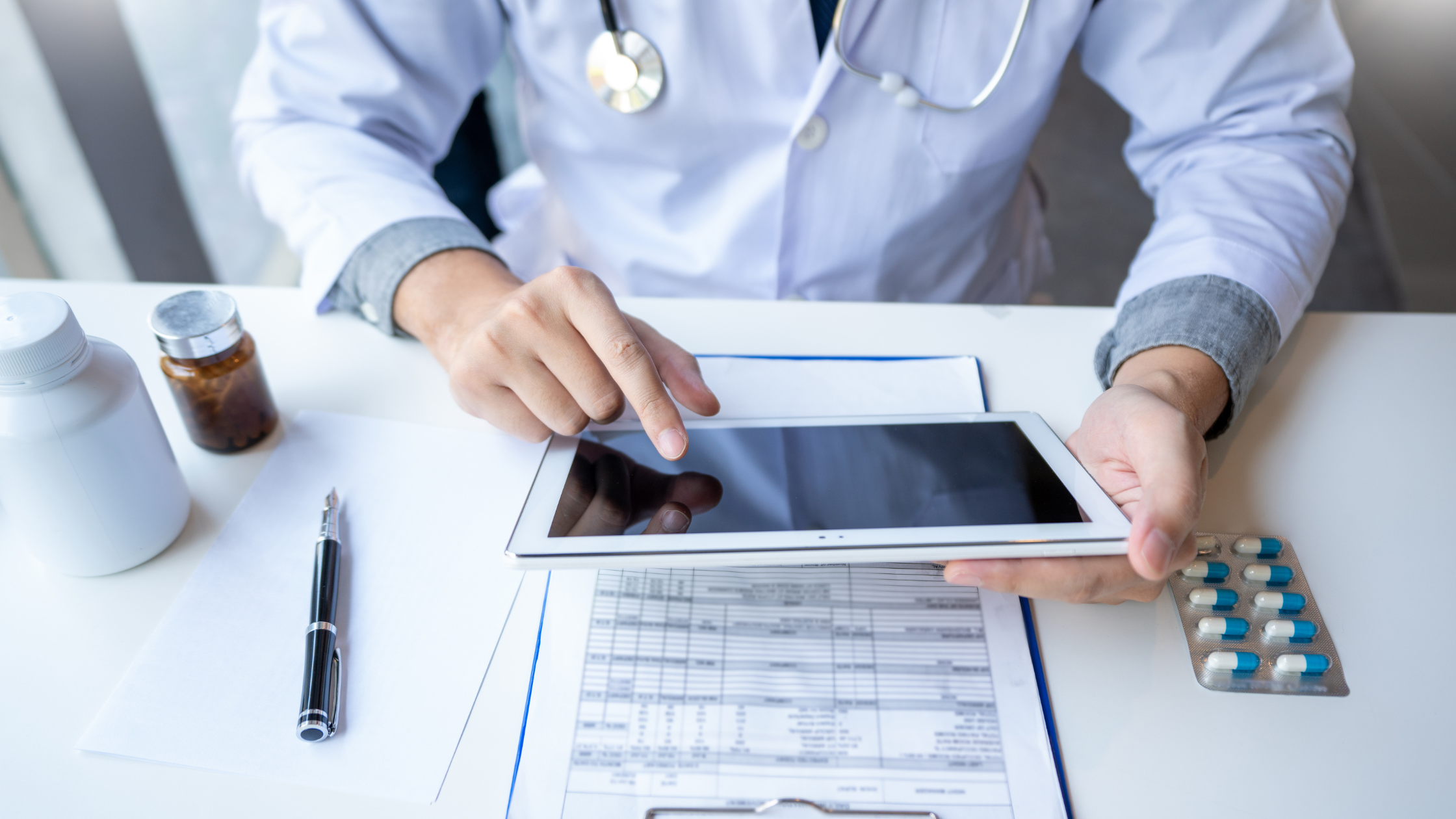 Image of a doctors hands holding a tablet using technology instead of paper. On the table a pen, pills and a clipboard.