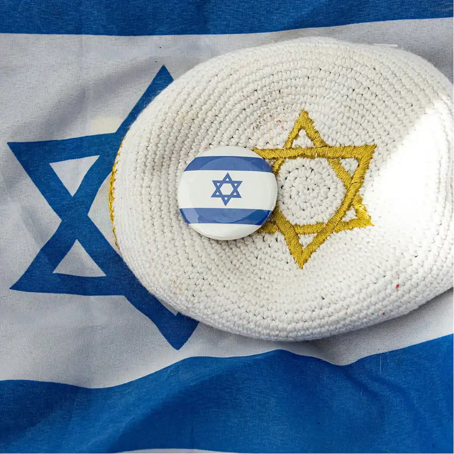 A white Kippah with an embroidered gold Star of David resting on an Israeli flag