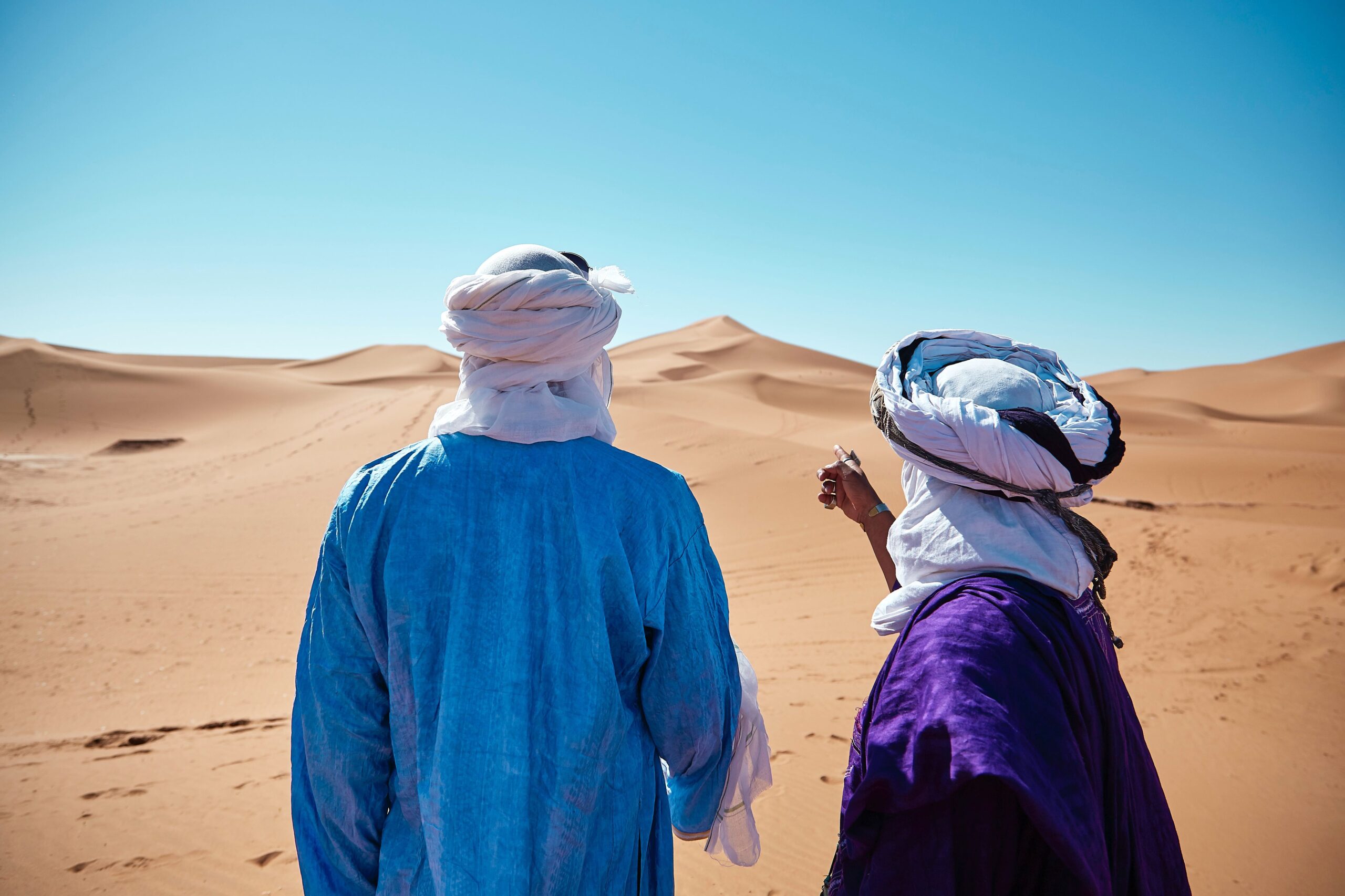Two people in colorful attire stand in a desert, one pointing at distant sand dunes under a clear blue sky, conveying exploration.