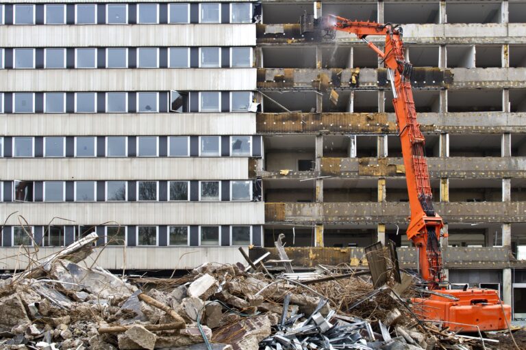 An orange excavator demolishes a building, revealing empty floors. Debris fills the foreground. The scene is industrial and dynamic.