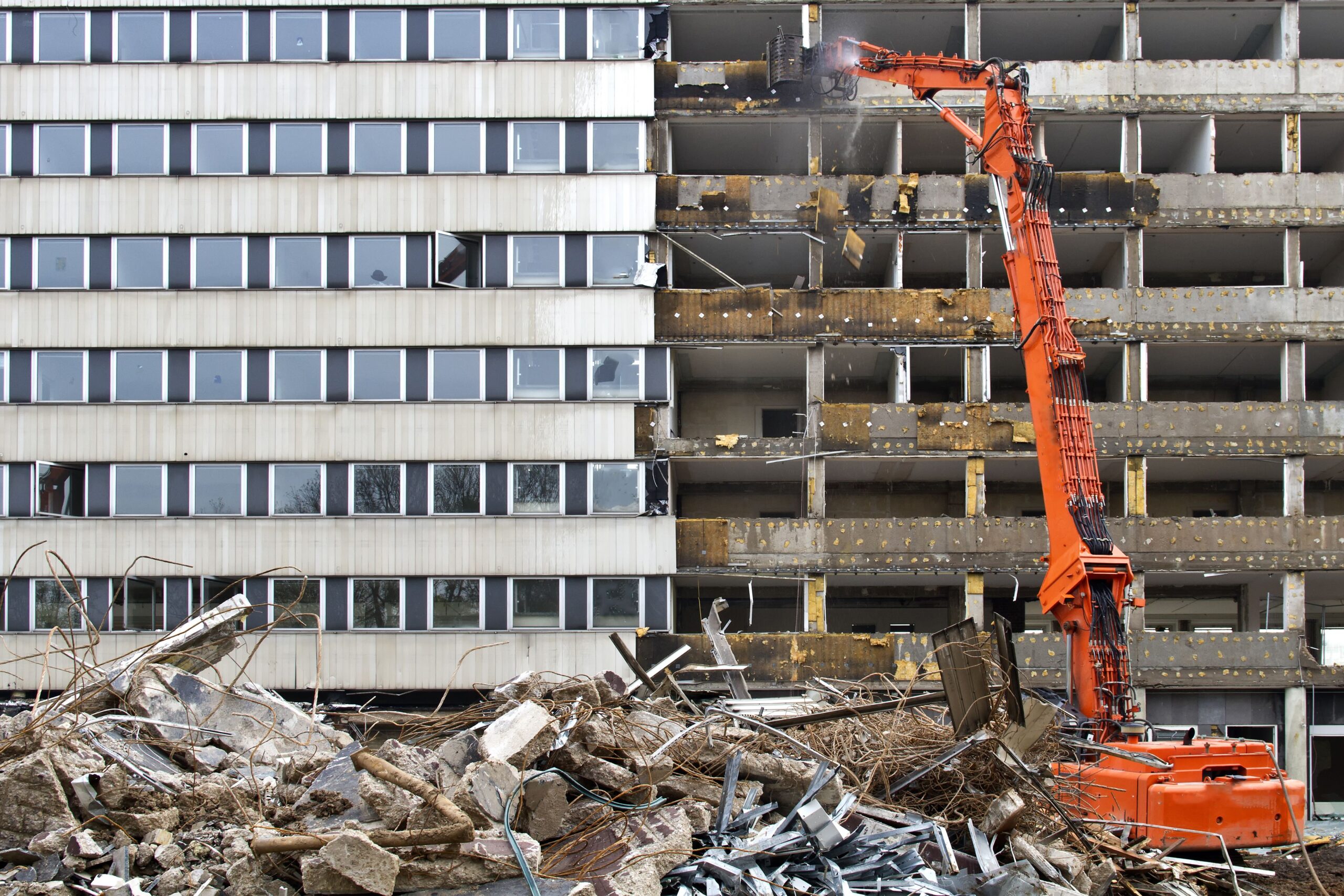 An orange excavator demolishes a building, revealing empty floors. Debris fills the foreground. The scene is industrial and dynamic.